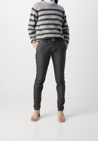 Person som bär svarta slim-fit byxor, grå och svarta randiga tröja, samt beige klacksandaler med korsade remmar, står med händerna i fickorna.