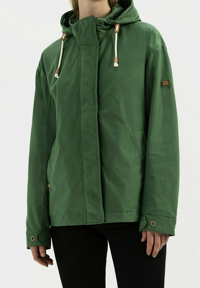 Veste à capuche verte avec cordons blancs portée par une personne en pantalon noir, fermeture éclair avant recouverte d'un rabat avec poignets à boutons-pression.