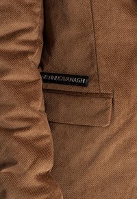 Chaqueta de pana marrón con un bolsillo en el lado izquierdo y una etiqueta con "Gráinne Kavanagh" en negro, con detalles de costura visibles en toda la prenda.