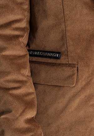 Hnědá manšestrová látka s klopovou kapsou a černou obdélníkovou štítkem s nápisem "JANNI KAVANAGH" přišitým nad kapsou.