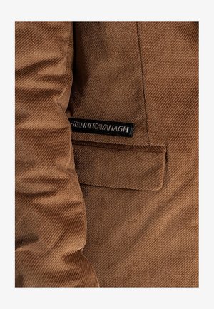 Tissu en velours côtelé marron avec une poche à rabat et une étiquette rectangulaire noire cousue au-dessus de la poche indiquant "JANNI KAVANAGH".