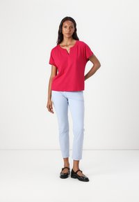Fuchsia Kurzarmoberteil mit V-Ausschnitt, kombiniert mit hellblauen, schmal geschnittenen Hosen und schwarzen Lackslippern. Einfaches und modernes Design.