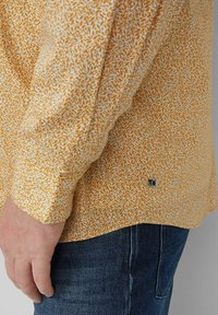 Pomarańczowa koszula z długim rękawem w drobny, biały wzór kwiatowy. Posiada zaokrąglony dół i metkę z logo przy dolnym szwie.