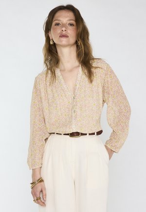 Woman wearing a light pink floral button-up blouse with cream high-waist pants, brown belt, gold hoop earrings, and bracelets against white background.