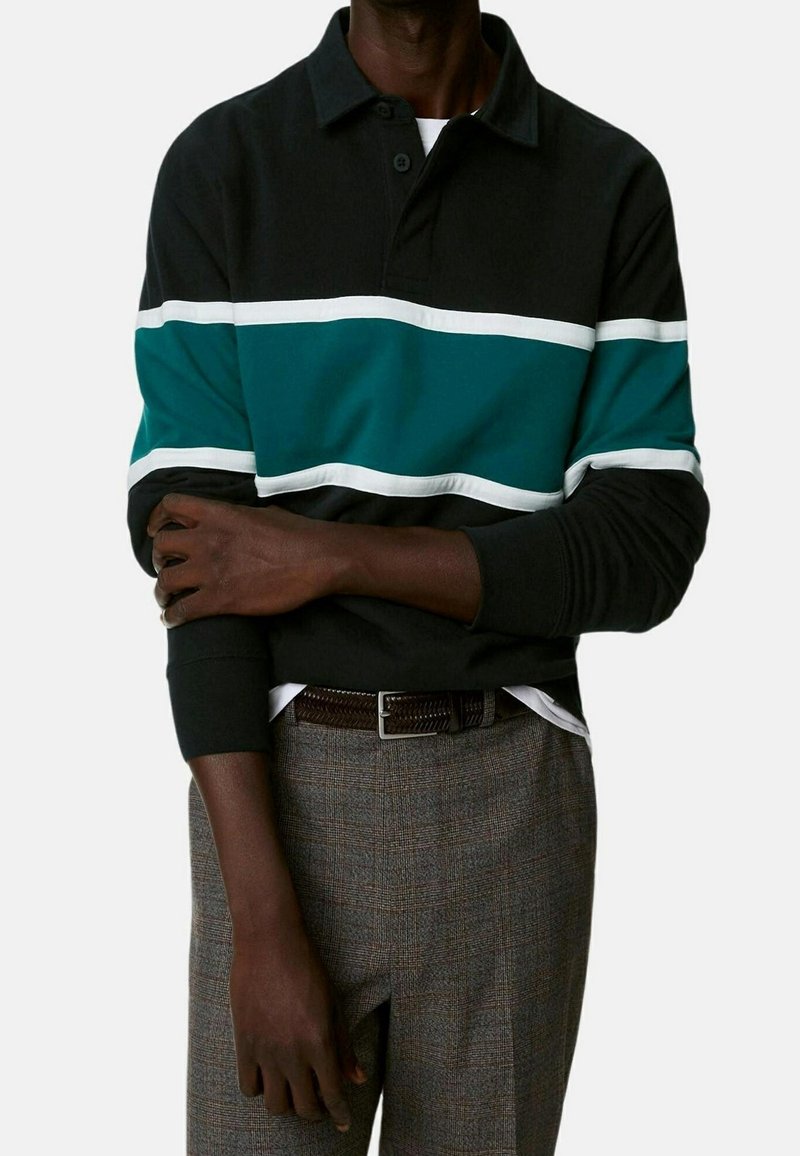 Homme portant un polo noir à manches longues avec des rayures horizontales vertes et blanches, un pantalon gris à carreaux, et une ceinture tressée marron, bras croisés.