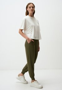 Witte t-shirt met korte mouwen gecombineerd met olijfgroene joggingsbroek met elastische boorden en zijzakken. Model draagt witte sneakers.