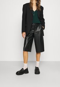 Blazer negro de gran tamaño, suéter verde texturizado, pantalones cortos de imitación cuero negros y mocasines negros de plataforma con calcetines a rayas.