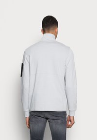 Ljust grå pullovermantel med hög krage, långa ärmar och svarta armbågsflekningar. Mjuk textur med minimala sömmar, designad för avslappnad användning.