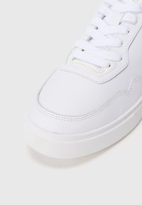 Tenis alb cu un superior din piele netedă, talpă din cauciuc texturat și șireturi albe plate. Detalii subtile de cusătură pe margini.