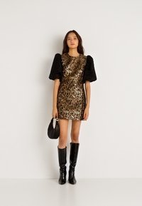 Mini φόρεμα λεοπάρ με λαμπερό ύφασμα, μαύρες πούφ μανσέτες, συνδυασμένο με μαύρες μπότες μέχρι το γόνατο και μια μικρή μαύρη τσάντα.