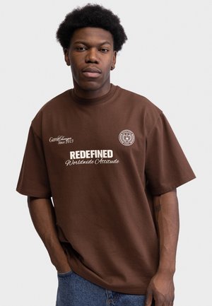 Giovane uomo con acconciatura afro che indossa una maglietta marrone con scritta bianca "REDEFINED Worldwide Attitude" e jeans blu, in piedi davanti a uno sfondo semplice.