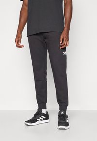 Črne trenirke s zožanim krojem, z elastičnim pasom, manšetami na gležnjih in belim Adidasovim logotipom na levem nogavici. Črne športne čevlje.