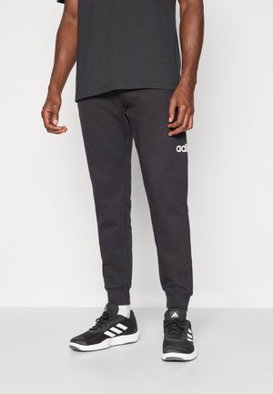 Schwarze Sweatpants mit schmaler Passform, ausgestattet mit einem elastischen Taillenbund, Bündchen an den Knöcheln und einem weißen Adidas-Logo am linken Bein. Schwarze Sportschuhe.