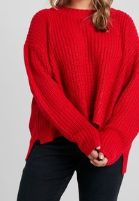 Czerwony sweter z dzianiny o luźnym kroju, z prążkowaną fakturą, szerokimi rękawami i bocznymi rozcięciami. W połączeniu z ciemnymi spodniami.