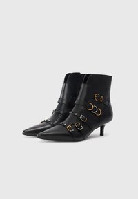 Botas de tacón medio de cuero negro con punta afilada, acentos de hebilla en oro y plata, y un diseño elegante. Textura suave.