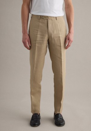 Homme portant un pantalon beige sur mesure, un t-shirt blanc et des mocassins noirs, debout sur un fond uni.