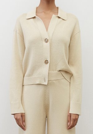 Frau trägt beige gestrickte Strickjacke mit zwei großen Knöpfen und passende hoch taillierte Strickhose vor schlichtem Hintergrund.