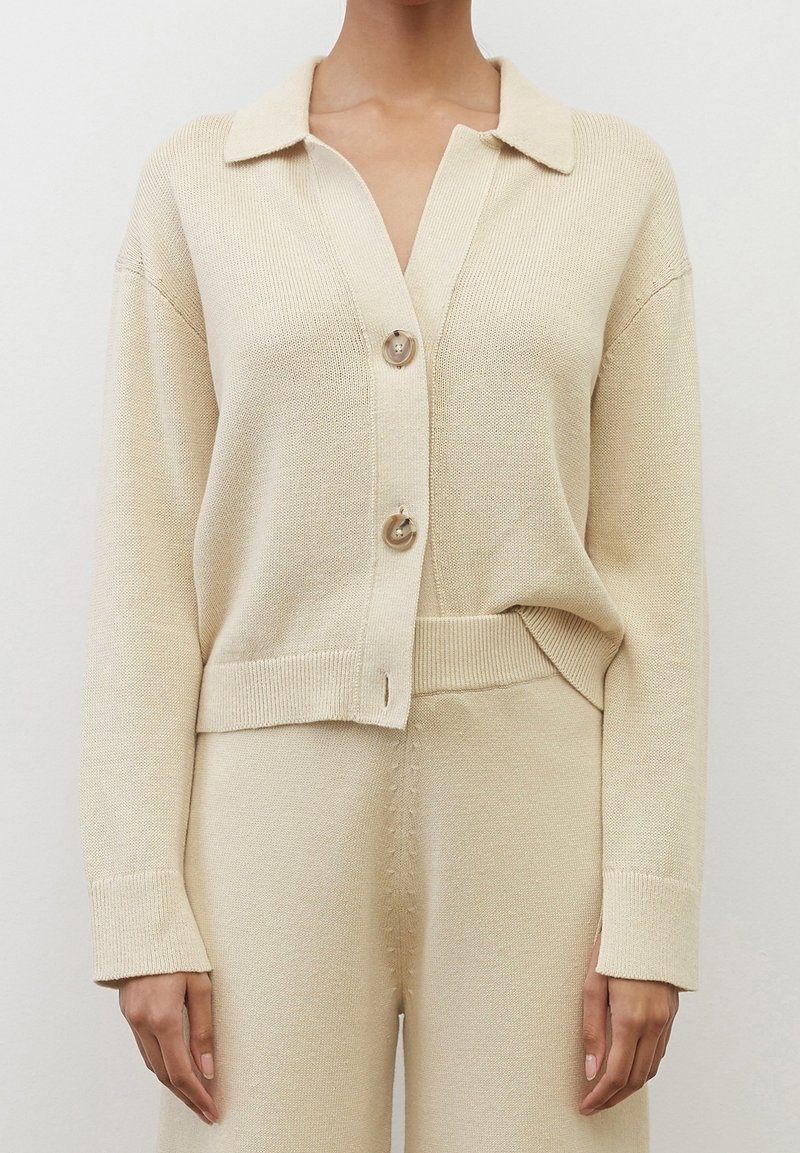 Femme portant un cardigan en tricot beige avec deux grands boutons et un pantalon en tricot taille haute assorti, sur un fond uni.