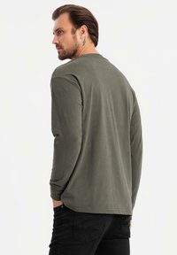Camicia a maniche lunghe verde oliva realizzata in morbido tessuto, con scollo rotondo e vestibilità comoda, abbinata a jeans neri.