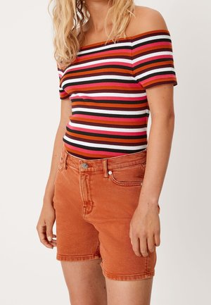 Femme portant un haut rayé épaules dénudées et un short en jean taille haute couleur rouille, debout devant un fond neutre.
