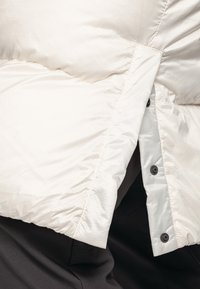 Pufferjack in licht crème, met een gladde, glanzende stof en een doorgestikt uiterlijk, gesloten met donkere knopen aan de voorkant.