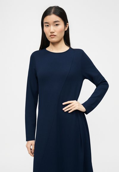 Νεαρή γυναίκα με μακριά μαύρα μαλλιά φοράει ένα μπλε σκούρο φόρεμα με μακριά μανίκια, στέκεται με το ένα χέρι στη μέση της μπροστά σε λευκό, απλό φόντο.