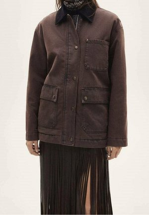 Persoon draagt een donkerbruine jasje met knopen en drie voorzakken, over een zwarte geplooide rok met een hoge, dijbeen-split.