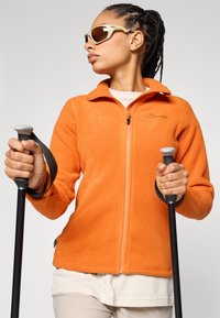 Orangene Fleecejacke mit Reißverschluss vorne, zwei Seitentaschen und Logo. Das Model hält schwarze Trekkingstöcke und trägt ein cremefarbenes Oberteil sowie eine Sonnenbrille.