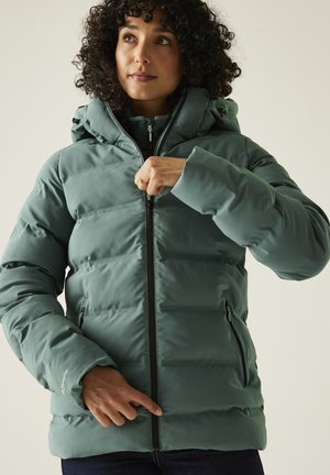 Frau mit lockigem Haar trägt eine grüne Steppjacke mit Kapuze und zieht den Reißverschluss hoch, vor einem schlichten hellen Hintergrund.