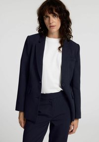 Marineblå skreddersydd blazer med en enkelt knappelukking, innsnitt lapeler og to frontlommer, stylet over en hvit t-skjorte.