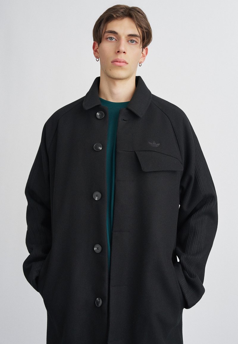 Cappotto nero con bottoni e colletto, dotato di tasca sul petto, maniche a costine e una texture liscia. Indossato sopra una maglietta verde petrolio scuro.