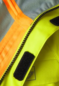 Dettaglio di giacca ad alta visibilità con tessuto giallo fluo, accenti arancioni, una cerniera nera e una patch in velcro all'interno.