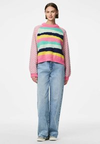 Pull en tricot multicolore avec des rayures roses, bleues, jaunes et marine, avec des manches roses texturées. Porté avec un jeans bleu clair.