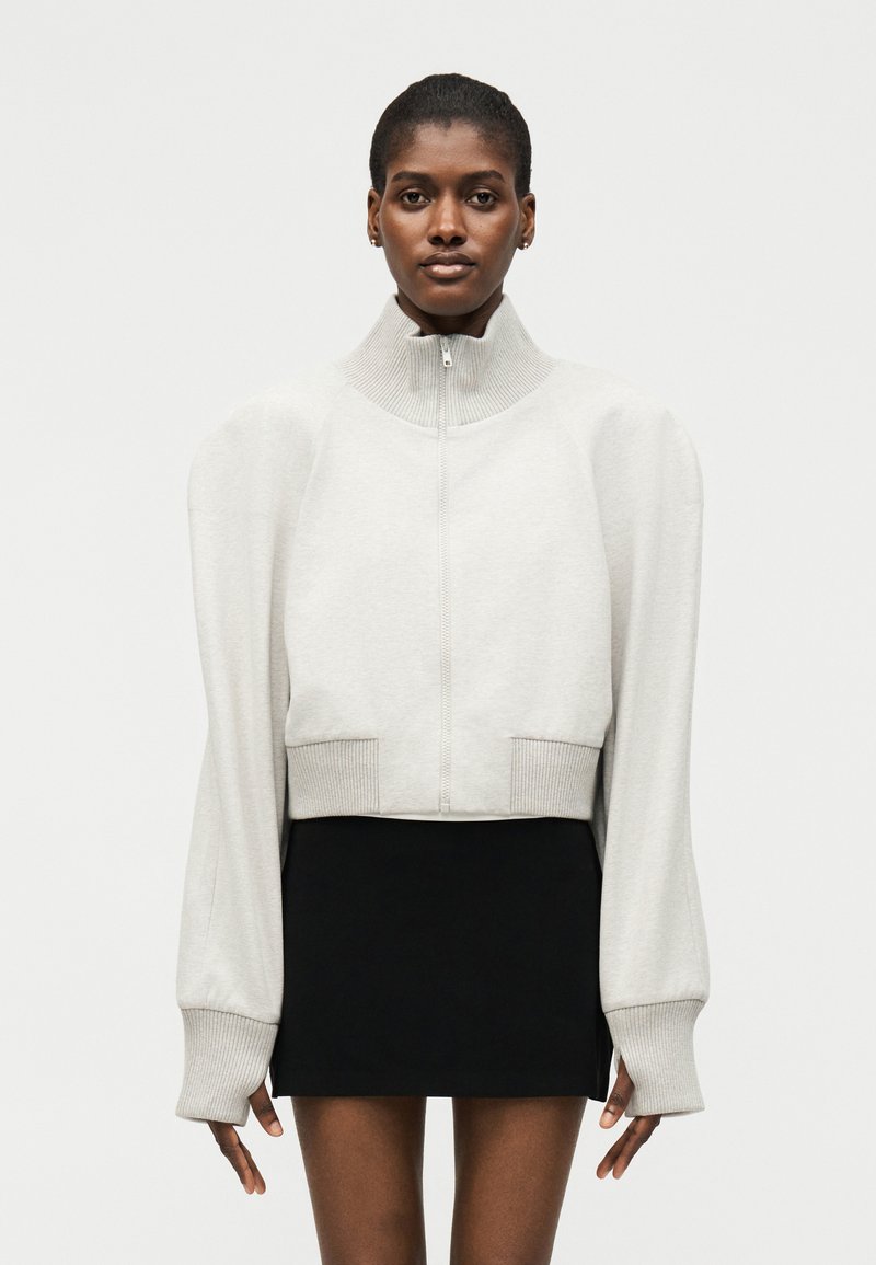 Γυναίκα με ανοιχτό γκρι πουλόβερ με φερμουάρ και φουσκωτά μανίκια και μαύρη μίνι φούστα, που στέκεται μπροστά σε ένα απλό ανοιχτό φόντο.