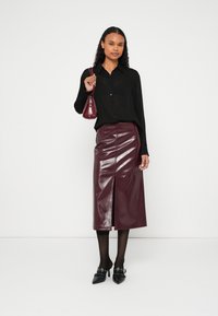 Μακριά, μπορντό φούστα από δέρμα με έντονη γυαλάδα και σχίσιμο μπροστά, σε συνδυασμό με μαύρο πουκάμισο με κουμπιά και μαύρα παπούτσια με τακούνι. Μικρή κόκκινη τσάντα ως αξεσουάρ.