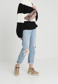 Persona che indossa un maglione a strisce bianche e nere, jeans chiari strappati, sneakers chunky beige e una borsa a tracolla rosa con paillettes.