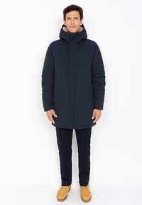 Abrigo largo acolchado azul marino con capucha, cierre de cremallera y dos bolsillos laterales. Lleva puesta unos pantalones negros y zapatos color marrón claro. Tela texturizada.