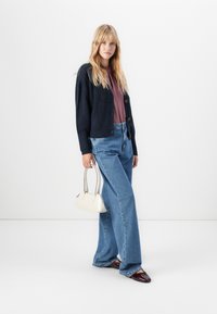 Femme blonde portant un haut violet, un cardigan marine, un jean bleu large et des chaussures bordeaux, tenant un petit sac à main blanc.