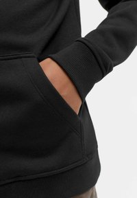Felpa nera con tasca a canguro, polsini e vita a coste. Realizzata in un tessuto morbido e strutturato con cuciture minime.
