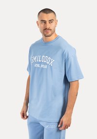 Helles blaues Baumwoll-T-Shirt mit kurzen Ärmeln, runder Halslinie und auffälligem weißen Text "SMILODOX ATHL. WEAR" auf der Vorderseite bedruckt.