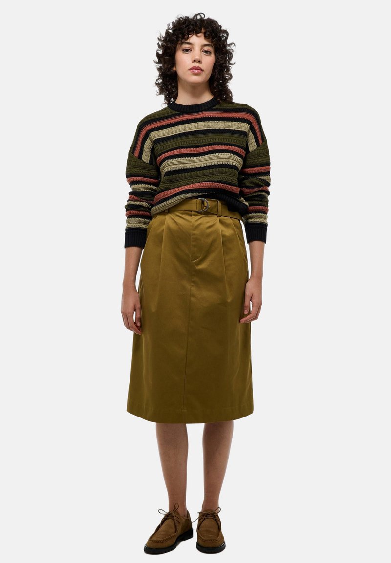 Femme aux cheveux bouclés, portant un pull rayé, une jupe ceinturée moutarde longueur genou et des chaussures lacées marron, se tenant devant un fond uni.