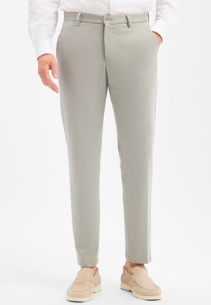 Mann trägt hellgraue Slim-Fit-Hose, weißes Hemd und beige Slipper, steht mit einer Hand in der Tasche.