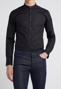 Camicia nera a maniche lunghe con collo alla coreana, caratterizzata da una chiusura con bottoni sul davanti e realizzata in tessuto liscio e leggero, abbinata a jeans blu scuro.