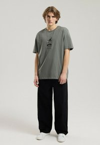 Jednolity oliwkowy T-shirt wykonany z bawełny, z okrągłym dekoltem i krótkimi rękawami, z nadrukiem z przodu. Zestawiony z czarnymi spodniami o szerokim kroju.