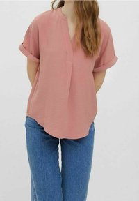 Blouse rose à manches courtes avec un col rond et une légère patte de boutonnage à l'avant. Le tissu est léger avec une finition texturée. Portée avec un jean bleu.