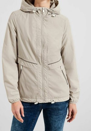 Veste à capuche beige avec fermeture éclair frontale, poches zippées et cordons ajustables, portée avec un jean bleu.