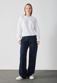 Pull blanc côtelé avec des épaules tombantes, associé à un pantalon en denim bleu foncé à larges jambes et des baskets blanches sur un sol gris.