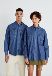 Denimskjorter for voksne, med lange ermer, to brystlommer og gult broderi, kombinert med lyse beige og olivengrønne bukser.