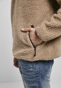 Tan fleecejakke med struktureret, blødt stof og sort lynlåslomme, parret med blå slidte jeans.