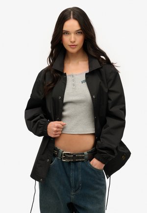 Sort, let jakke med høj krave, trykknaplukning, elastiske mansjetter og snoretræk i kanten, båret over en grå cropped tanktop og blå jeans.